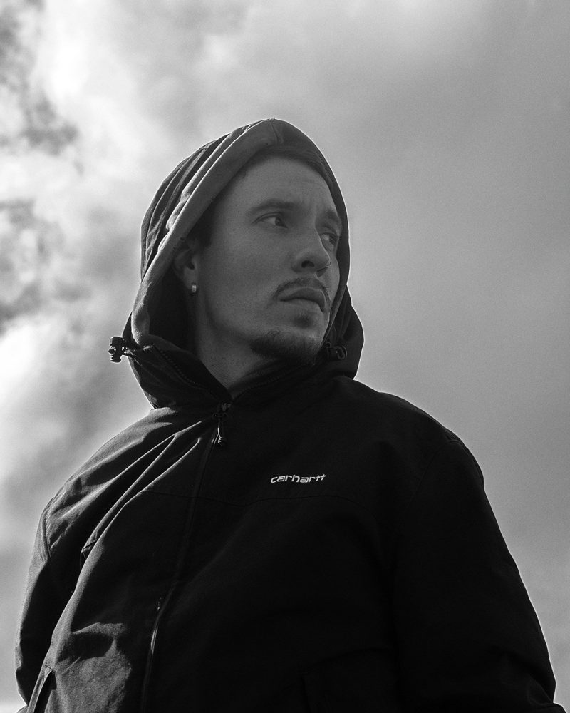 Schwarzweiß-Portrait eines Mannes mit Kapuze vor Wolkenhimmel